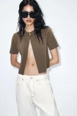 Chaqueta corta color verde oliva, de tejido de punto con cuello redondo y manga corta. Cierre frontal con un único botón dorado.