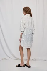 Vestido camisero corto y de silueta amplia en color blanco, con cuello clásico, cierre frontal de botones y mangas largas. Cuenta con diseño bordado floral en toda la prenda y dobladillo asimétrico, más largo en la parte trasera.