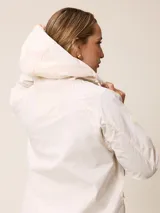 Campera de corte recto y estilo funcional, color beige, con capucha integrada, cierre frontal con botones a presión y bolsillos delanteros amplios. Presenta un diseño minimalista con cordones ajustables en la capucha.