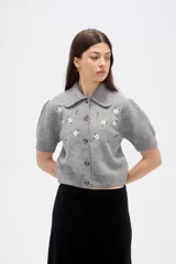 Saco de punto gris con cuello tipo camisa, manga corta y cierre frontal con botones. Presenta un delicado bordado de flores blancas en la parte superior delantera.