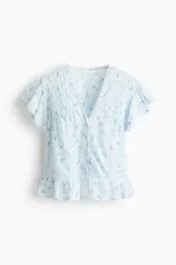 Blusa celeste claro con delicado estampado floretado azul, escote en V pronunciado, mangas cortas tipo mariposa y cierre frontal con botones. Presenta inserciones de encaje y alforzas decorativas en la parte delantera, además de un dobladillo con volados y detalles de encaje.