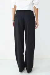 Pantalón negro de tiro medio con pliegues frontales, confeccionado en tejido sastrero fluido. Presenta piernas amplias, bolsillos laterales y cierre frontal con cremallera y botón.