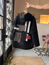 Campera con capucha de textura tipo corderito en color negro, con un diseño de collage geométrico en la espalda que combina parches en tonos marrón, negro, blanco y rojo. Presenta un corte holgado y diseño artesanal único.