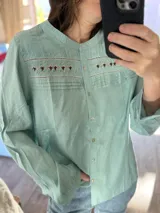 Blusa de manga larga color blanco con cuello redondo y cierre frontal de botones. Presenta detalles de bordado estilo punto smock en el pecho y textura de rayas verticales en la parte superior. Diseño holgado y estilo bohemio.