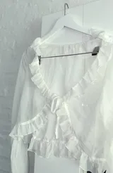 Blusa blanca traslúcida con escote en V, volados en el contorno y mangas largas acampanadas con lazos en los puños.