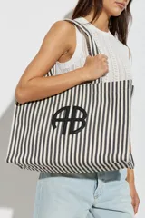 Bolso tipo shopper de diseño rectangular y amplio, confeccionado en tela con patrón de rayas verticales en color blanco y negro. Cuenta con dos asas superiores para llevar al hombro y detalle de aplique metálico con monograma en el centro.