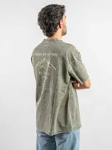 Remera de corte oversize color verde con efecto lavado, manga corta y cuello redondo. Presenta un estampado gráfico en la espalda con el diseño de una montaña y la frase 'Fresh air is free, don't be afraid of the high'.