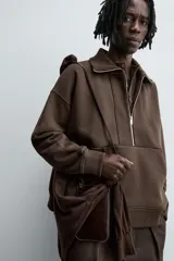 Sudadera de corte relajado color marrón oscuro, con cuello subido y cierre frontal de cremallera. Presenta manga larga, bolsillo frontal tipo canguro y acabados en rib en puños y ruedo.