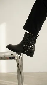 Bota de caña alta color negro, con diseño versátil que incluye un arnés desmontable con hebilla y argollas metálicas. Presenta costuras decorativas en la parte superior de la caña y un taco bajo cuadrado.