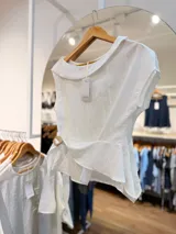 Blusa blanca de diseño cruzado sin mangas, con escote bote y un lazo lateral con hebilla para ajustar en la cintura.