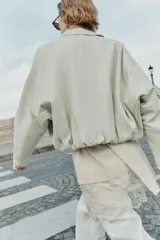 Chaqueta estilo bomber de cuero sintético en color beige, con cuello solapa, cierre frontal con cremallera, mangas largas y bajo elástico fruncido.