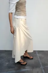 Falda midi de tiro medio color gris, con diseño asimétrico en el bajo, bolsillos frontales y traseros, y cierre frontal con cremallera y botón.