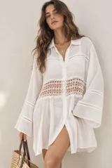 Vestido corto beige estilo camisa con mangas largas amplias, cuello camisero, cierre frontal con botones y detalles de encaje calado en el pecho y las mangas.