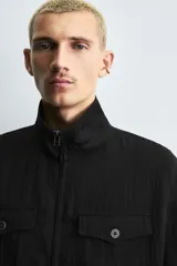 Cazadora de hombre color verde oliva, estilo bomber o Harrington, con cuello subido, cierre frontal de cremallera y puños y bajo elásticos. Presenta bolsillos de plastrón con solapa en el pecho y bolsillos de vivo en la cadera.