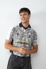 Camiseta de estilo fútbol con estampado de camuflaje gris, cuello tipo polo negro y mangas blancas. Presenta gráficos frontales con la palabra LEGACY en amarillo y detalles de escudos deportivos.