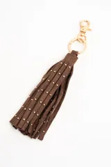 Charm para bolso compuesto por flecos de cuero sintético color marrón, decorados con pequeñas tachas metálicas doradas. Incluye un aro y un cierre de mosquetón metálico en tono dorado.