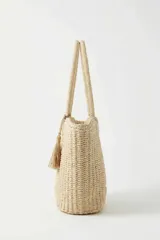 Cartera tipo shopper de mimbre tejido color beige, con doble asa de hombro y detalle de borla colgante.