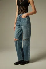 Pantalon de jean azul con corte recto y ancho, presenta aberturas en las rodillas y detalles de tachas metálicas a lo largo de las costuras laterales.