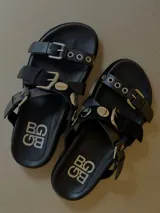 Sandalias planas de cuero color negro con triple tira, hebillas metálicas, ojales decorativos y tachas.