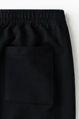 Jogger de punto de algodón color negro, con cintura elástica y cordón de ajuste, bolsillos laterales y pliegues frontales pespunteados. Corte relajado de pernera recta.