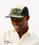 Gorra tipo 5 panel de microfibra con diseño de bloques de colores en beige, verde oliva y negro. Presenta visera recta y una etiqueta frontal bordada con la frase Stay Grounded y un gráfico de montaña y globo terráqueo. Incluye cierre posterior ajustable.