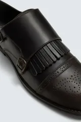 Zapato de vestir estilo monk strap fabricado en piel color marrón oscuro. Presenta ajuste mediante dos hebillas laterales metálicas, detalle de flecos en el empeine y puntera con perforaciones decorativas tipo brogue.