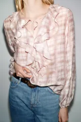 Blusa de manga larga abullonada con puños elásticos, cuello redondo con volantes y detalle de cordón ajustable en el escote pico. Presenta un estampado a cuadros en tonos blanco y rosado/marrón claro.