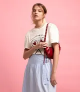 Cartera tipo shoulder bag de diseño media luna, confeccionada en poliuretano con acabado brillante. Presenta detalles frontales estilo gota, cierre superior por cremallera y correa de hombro ajustable con ojetes metálicos.