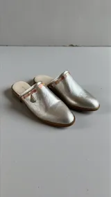 Sandalias chatitas tipo mules, color plateado con detalle de arte en la parte superior y aplique metálico triangular.