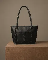 Bolso tipo tote de cuero vacuno negro, hecho a mano, con dos asas de hombro. Presenta un bolsillo exterior frontal con costura decorativa en forma de espiga y detalles de costura trenzada en los laterales. Incluye un dije de borla de cuero colgando de una de las asas.