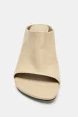 Sandalia tipo mule con tacón cuña, confeccionada en piel color beige. Presenta diseño destalonado, apertura frontal y punta cuadrada asimétrica.