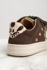 Zapatillas bajas de gamuza color marrón con cordones y suela de goma color crema. Presentan una franja lateral con estampado de vaca y el logo de la marca bordado en el lateral.