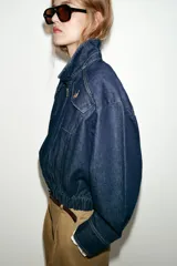 Chaqueta estilo bomber confeccionada en tejido denim azul oscuro. Presenta cuello solapa, cierre frontal con cremallera, mangas largas y bajo elástico. Incluye bolsillos delanteros de plastrón con solapa y botones.