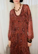 Vestido largo de corte evasé y diseño oversize, confeccionado en tejido semitransparente con estampado paisley en tonos rojizos y marrones. Presenta cuello escotado con cierre delantero, mangas largas con puños abullonados y forro interior a tono.