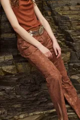 Pantalón de cuero color marrón óxido con acabado brillante o texturizado. Diseño de tiro alto con corte recto y abertura en el tobillo. Se complementa con un cinturón ancho de cuero marrón con hebilla grande y detalles metálicos plateados.