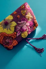 Bolso tipo sobre color violeta con diseño de abalorios y cuentas bordadas formando un patrón floral multicolor. Cuenta con una borla colgante en el cierre y correa de cadena extraíble.