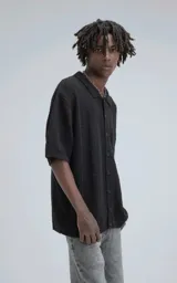 Camisa de manga corta tejida en punto abierto negro, con cuello polo y cierre frontal de botones. Presenta un calce oversize.