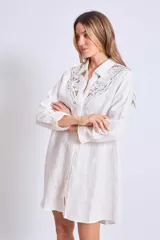 Vestido camisero corto color crudo, de lino, con cuello camisero, mangas largas y cierre frontal con botones. Presenta apliques de encaje en cuello y hombros.