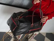 Bolso weekender de cuero negro, con cierres metálicos rojos, asas de mano y correa ajustable para hombro en tono marrón.
