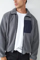 Campera de tejido polar tipo teddy en color gris, con cuello camisero, cierre frontal de cremallera y bolsillo funcional en el pecho con solapa de contraste.