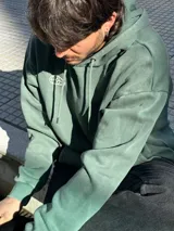 Canguro verde con capucha y cordones ajustables. Presenta un bolsillo tipo canguro y dos estampados blancos: un logo con forma de globo terráqueo en el lado izquierdo del pecho y una flor en el lado derecho.