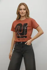 Remera de algodón color terracota con cuello redondo y mangas cortas. Presenta un diseño gráfico frontal con aplicación de lentejuelas negras y texto estampado.