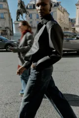 Cazadora corta negra de efecto piel, con cuello solapa, manga larga y cierre frontal con botones a presión ocultos. Incluye bolsillos delanteros de vivo con botón a presión y bajo con elástico en la espalda.