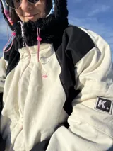 Campera oversize de gabardina combinada en colores crudo y negro, con cierre frontal y capucha de piel sintética negra. El interior está forrado en polar con diseño estampado.