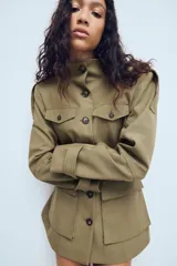 Chaqueta de estilo militar en color beige, con cuello subido, cierre frontal de botones, bolsillos de plastrón con solapa en el frente y detalle de hombreras con trabillas.