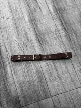 Cinto marrón de cuero con hebilla metálica plateada y ojales metálicos.