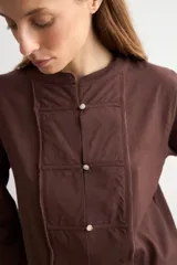 Blusa de manga larga color marrón con cuello redondo y cierre frontal con alamares. Presenta costuras visibles y diseño estructurado.