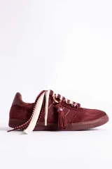 Zapatillas urbanas color tostado con textura geométrica en el cuerpo y detalles de gamuza en la puntera y laterales. Presentan un diseño distintivo con cordones anchos tejidos en tonos tostado y blanco, y un accesorio metálico con borla de gamuza en el empeine.