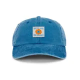 Gorro de visera curva, estilo dad hat, color azul lavado con efecto desgastado. Presenta un parche rectangular bordado al frente con un diseño de flor naranja y azul y el texto "RHYTHM LIVIN" y "SURF ESSENTIALS".