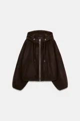 Chaqueta estilo bomber de corte oversize en color marrón oscuro, confeccionada en tejido suave. Presenta capucha con cordones ajustables, cierre frontal con cremallera, mangas largas con puños elásticos y bolsillos delanteros de vivo. El bajo cuenta con detalle de pliegues.